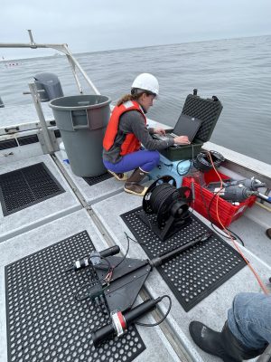 A scientist onboard a small boat working at a laptop, sitting next to a black instrument on the floor of the boat