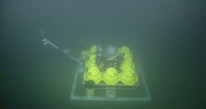 A silver metal sphere surrounded by eight yellow plastic spheres and forming a three-by-three grid on a square, metal frame. A metal arm with a cylinder-shaped, metal canister extends off the left. This screen capture from underwater video shows equipment sitting on greenish-brown mud in murky waters.