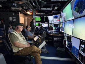 A scientist sits in the dark submersible control room aboard a research ship. He is operating camera controls while watching a wall of monitors displaying live video and data feeds from a robotic submersible exploring the deep seafloor. In the background are three submersible pilots and a scientist.
