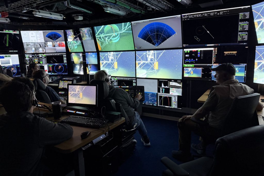 Two scientists and three submersible pilots sit in the dark submersible control room aboard a research ship. All have their backs to the camera as they watch a wall of monitors displaying live video and navigation data from the submersible as it installs a scientific instrument on the deep seafloor.