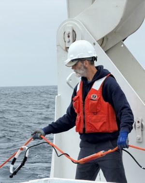 A scientist wearing a white hard hat, orange life vest, and blue gloves deploys a scientific instrument from the back deck of a research ship. He is holding an orange cable connected to a mooring outside of frame. In the background is gray-blue ocean, with overcast gray sky on the horizon.