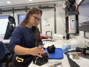 A scientist processes a rock sample in the lab aboard a research ship. She is removing pens from a black bag. The softball-sized black rock is on a blue tray on the counter in front of the scientist. In the background are the white walls of a brightly lit scientific lab.