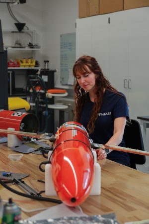 A researcher seated at a work bench disassembles a robotic glider inside a brightly lit engineering lab.