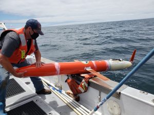 An engineer in a gray short-sleeved shirt, orange life vest, blue jeans, brown boots, blue cap, and black face mask prepares to deploy a bright orange, plane-shaped robot over the edge of a research ship. The robot is a glider with short orange “wings,” a torpedo-shaped orange body, and a white tail section. The robot is balanced on a life vest on the silver metal edge of a research ship. Four thin white plastic rods, a thin blue plastic rod, and two textured black mats are visible on the deck of the ship. Photographed outdoors with dark blue ocean and gray sky visible in the background.