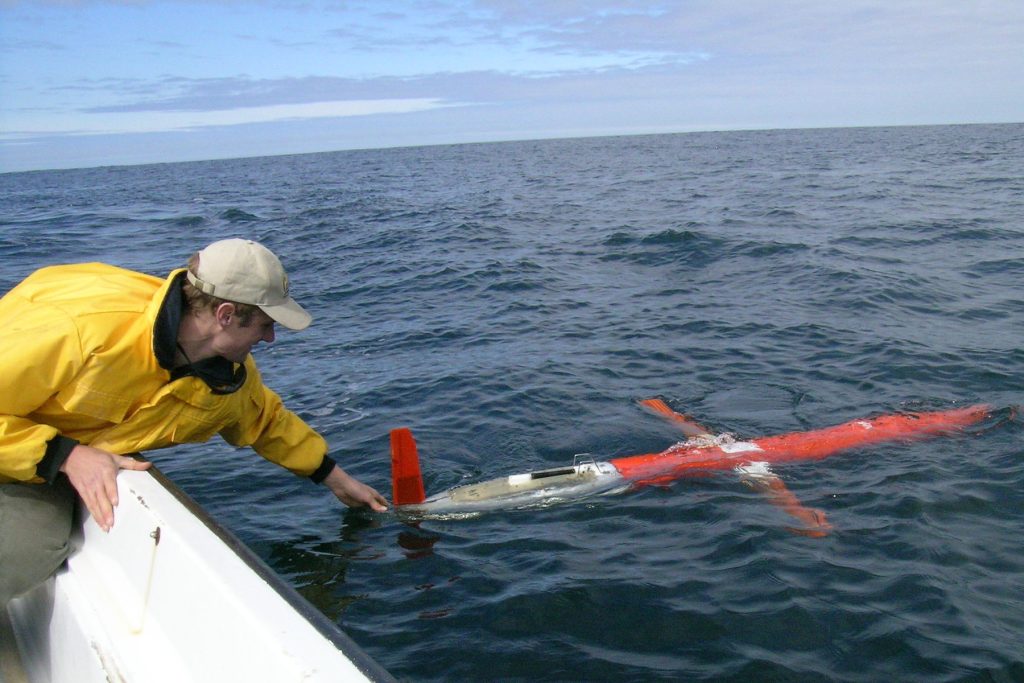 A research technician in a yellow jacket and brown pants sits on a white ship and examines a plane-shaped robot after deployment in the ocean. The robot is a glider with short orange “wings,” a torpedo-shaped orange body, and a white tail section. The technician was photographed outdoors on an overcast day with clouds and dark blue ocean in the background.