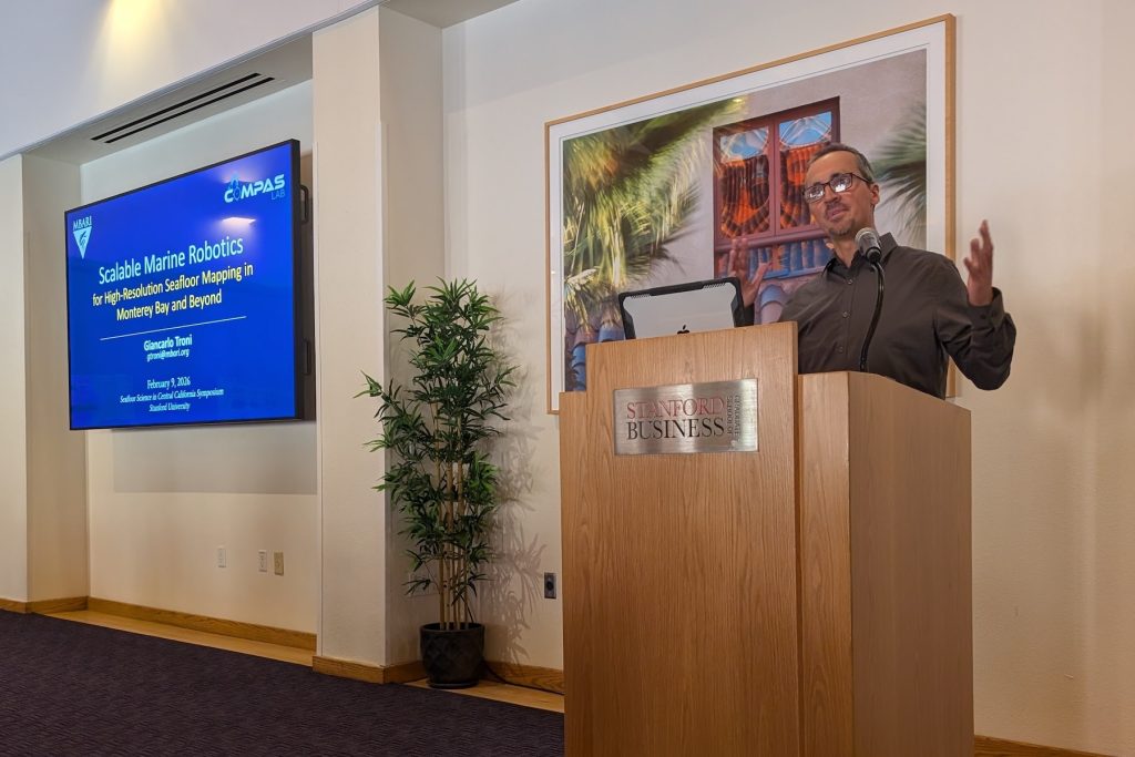 An engineer presents slides at a symposium. The engineer has short salt-and-pepper hair and a goatee, is wearing glasses and an olive-green shirt, and is standing behind a brown wooden podium with a silver metal plaque with the logo for the Stanford Graduate School of Business. On the left side of the frame is a blue slide with the MBARI and CoMPAS Lab logos and the title that reads, Scalable Marine Robotics. In the background are a large photo print, a green plant, and white walls.