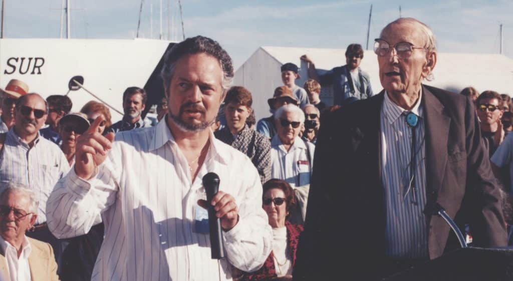 Steve Etchemendy in a white collared shirt holding a microphone (left) and David Packard in a black blazer, striped shirt, and silver bolo tie (right), standing in front of a crowd of people outdoors on MBARI’s dock.