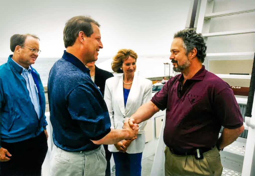 Steve Etchemendy in maroon polo shirt and khaki pants (right) shaking hands with Vice President Al Gore in a navy polo shirt and beige pants, with Representative Sam Farr in a teal jacket, blue shirt, and black pants (background left) and Marcia McNutt in a white blazer, blue shirt, and blue pants (background right) outdoors on the deck of an MBARI research vessel.