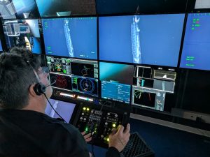 A scientist sitting in the dark submersible control room aboard a research ship watches a wall of monitors displaying live video and data feeds from a robotic submersible. He is operating a metal box with camera controls. The screens are displaying video of a deep-sea siphonophore with a gelatinous, rope-like body.