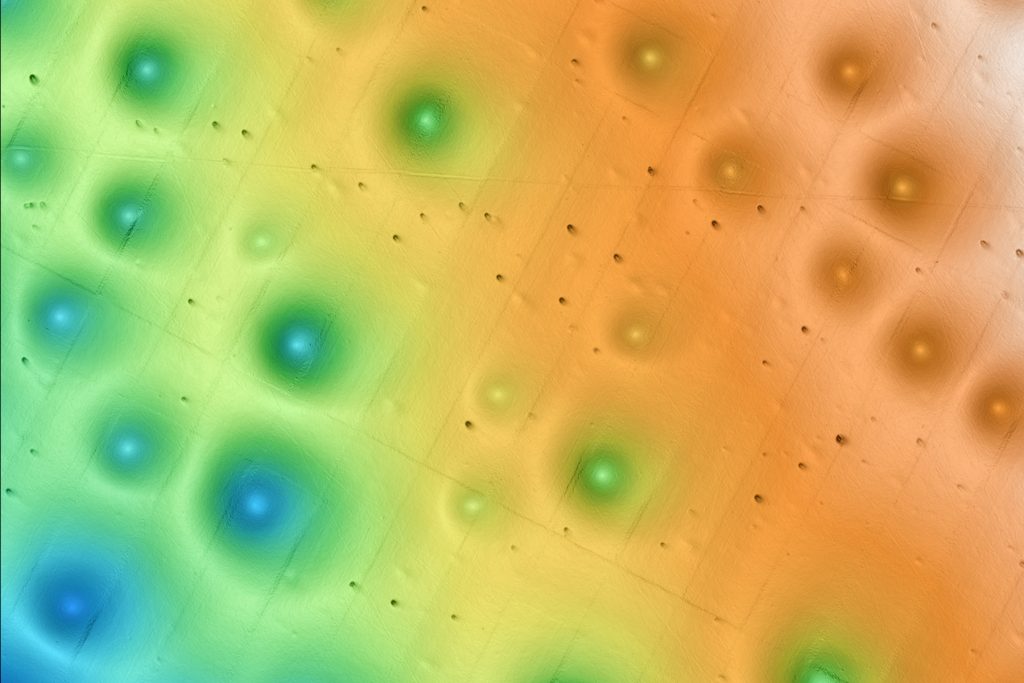 A sample of multi-colored seafloor bathymetry shows circular depressions called pockmarks on the seafloor. The image color represents a depth gradient with shallower depths in orange on the right transitioning to yellow, green, and blue on the left to represent deeper depths. The pockmark formations are circular in shape and spaced evenly apart. There are 18 large pockmark formations in the frame, as well as four smaller pockmark formations and a handful of other small, dark pits on the seafloor.