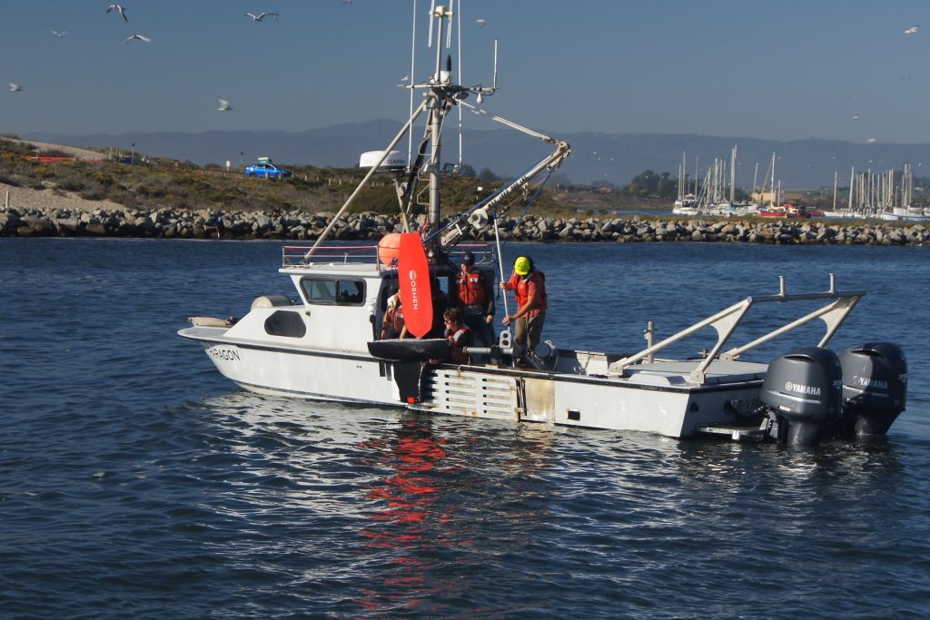 Marine operations crew deploy a scientific robot over the side of a white research boat. The crew are wearing hard hats and orange life vests. They are holding a robot with a black float and orange plastic sail over the water. The ship is drifting in coastal waters, with a jetty, harbor, and ship masts in the background and coastal mountains and hazy blue sky on the horizon.