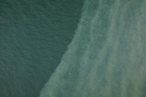 Aerial view of a color front formed by two bodies of water converging on the ocean’s surface. The seawater on the left is dark greenish-blue, while the seawater on the right has bands of fine, light-brown sediment. The color front bisects the image diagonally from the top right to bottom left.