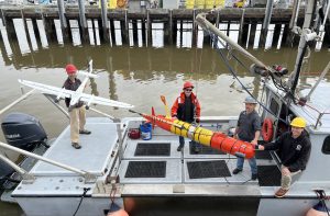 Four MBARI engineers pose for a photo on the silver metal deck of a research vessel while preparing for deployment of advanced robots for studying the ocean. On the left is an engineer wearing a red hard hat, gray jacket, beige pants, and gray shoes and holding a white plane-shaped drone. In the center a crane holds a suspended red-and-orange torpedo-shaped underwater robot. Standing around the robot are an engineer wearing a red hard hat, a black-and-orange jacket, blue jeans, and black shoes, an engineer wearing a gray hard hat, gray jacket, blue jeans, and black boots, and an engineer wearing a yellow hard hat, black hooded sweatshirt, beige pants, and brown boots. The brownish-green water of the harbor and pilings from the gray dock are visible in the background.