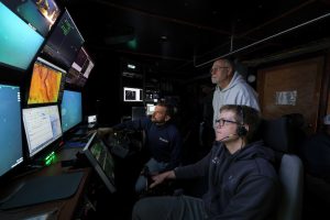 Two engineers and an ROV pilot watch video and mapping data displayed on a bank of video monitors in the dark control room of a research ship. The engineer on the left is seated, the engineer in the center is standing, and the ROV pilot is seated in the foreground with his right hand on a joystick.
