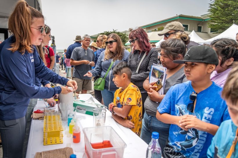 An MBARI researcher conducts a science demonstration for a large audience of Open House visitors. The MBARI researcher has shoulder-length brown hair, is wearing sunglasses, a navy blue pullover, and gray pants, and is standing on the left side of the frame. In the center is a table of test tubes and science activities. On the right side of the frame is a crowd of Open House visitors. The background is overcast gray sky and MBARI’s research buildings.