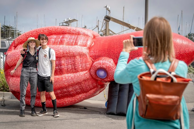Two visitors pose for a photo in front of a large, bright red inflatable model of a vampire squid. The visitor on the left is wearing a beige hat, black shirt, gray jeans, and black boots. The visitor on the right has shaggy black hair and is wearing a white shirt, black shorts, black socks, and black shoes. In the foreground on the right side of the frame is a third visitor with shoulder-length blonde hair wearing a green jacket and brown backpack, taking a photo. In the foreground is the asphalt of MBARI’s parking lot. In the background are the masts of ships in Moss Landing Harbor and an overcast gray sky.