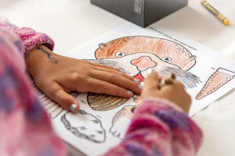 A young Open House visitor colors in an illustration of a sea otter with a brown crayon. The child has silver nail polish and is wearing a pink-and-purple fleece jacket. In the background is white butcher paper.