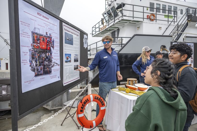 A member of MBARI’s ship crew speaks with a pair of Open House visitors. The crew member has shaggy brown hair and a beard and is wearing sunglasses, a navy blue pullover, and blue jeans. He is standing in the center of the frame, pointing to a poster identifying the components of a robotic submersible on the left side of the frame. The visitors are standing on the right side of the frame in the foreground. In the background are other members of MBARI’s ship crew and an MBARI research vessel.