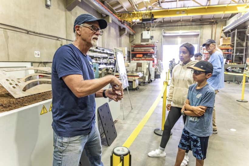An MBARI engineer speaks with two children while two adults observe in the distance. The engineer has a gray goatee, is wearing a navy blue cap, navy blue pullover, and blue jeans, and is standing on the left side of the frame behind a yellow stanchion. The children are on the right side of the frame. In the background is an assortment of machinery inside MBARI’s machine shop.