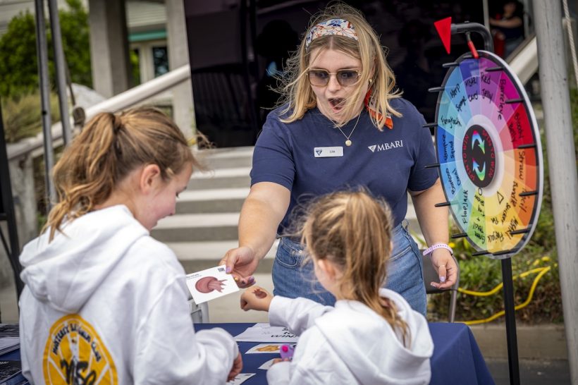 An MBARI staff member hands a sticker to a pair of Open House visitors. The staff member has shoulder-length blonde hair, is wearing a colorful headband, sunglasses, a navy blue t-shirt, and blue jeans, and is standing next to a multi-colored spin wheel. In the foreground are two children wearing white hooded sweatshirts. In the background are the steps and entrance to MBARI’s building.