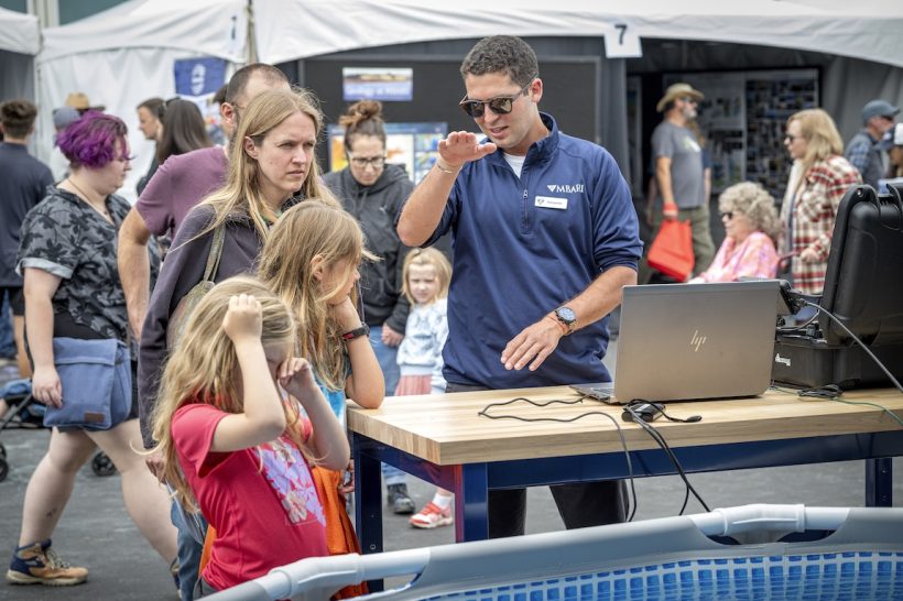 An MBARI engineer explains a robot to Open House visitors. The engineer has short black hair and is wearing sunglasses, a navy blue pullover, a watch, and gray pants. He is on the right side of the frame, standing behind a wooden counter with a laptop and black plastic case. On the left side of the frame are four visitors observing a pool with an underwater robot. In the background are tents and several visitors walking past.