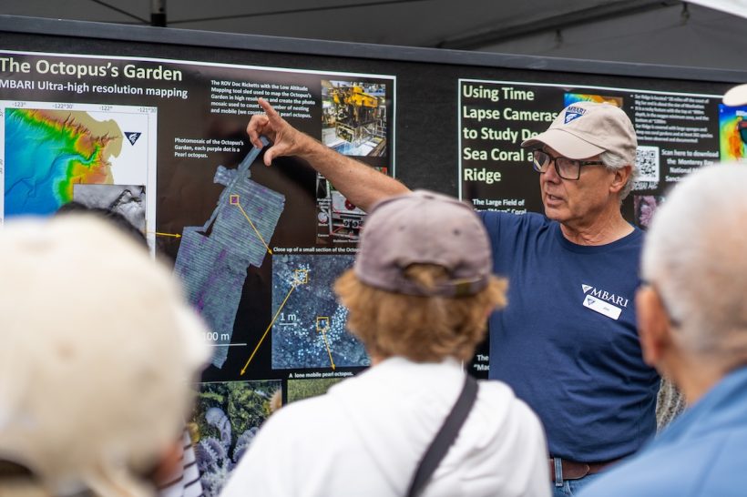 An MBARI researcher talks to a crowd of Open House visitors. The researcher is wearing a beige cap, glasses, a navy blue t-shirt, and blue jeans. He is standing in front of scientific posters describing MBARI research at the Octopus Garden and Sur Ridge. In the foreground are the backs of the heads of three visitors.