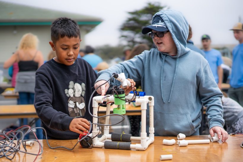 Two Open House visitors build an underwater robot. The child on the left is wearing a black graphic sweatshirt. The child on the right is wearing a blue cap, sunglasses, and a light blue hooded sweatshirt. They are standing behind a brown wooden table with an assortment of plastic parts assembled into the square frame of a robot. In the background are other Open House visitors blurred out of focus.