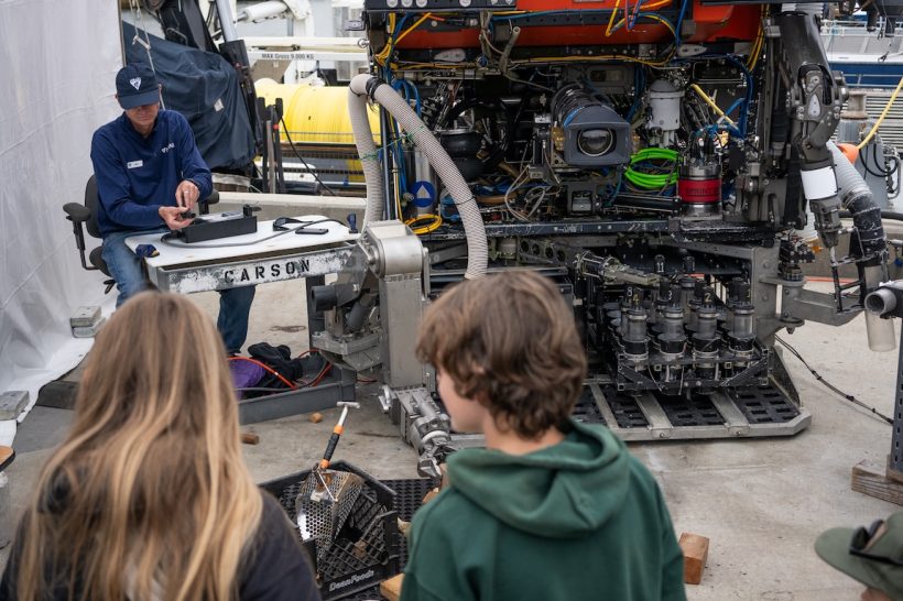 An MBARI ROV pilot controls the mechanical arm of a robotic submersible. The pilot is wearing a navy blue cap, navy blue pullover, and blue jeans, and is sitting on the left side of the frame. On the right side of the frame is a robotic submersible with an orange foam float and black metal scientific instrumentation. In the foreground are two children attending the MBARI Open House.