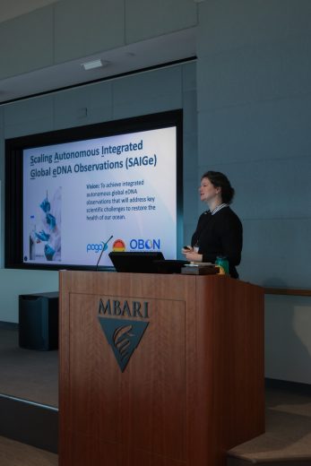 A scientist presents slides to an audience. The scientist has long black hair pulled back in a bun and is wearing a blue shirt and dark gray sweater. She is standing behind a wooden podium with the MBARI logo and holding a laser pointer. Behind her is a projection screen displaying a presentation slide titled, Scaling Autonomous Integrated Global eDNA Observations (SAIGe). In the background are the gray walls of a conference room.