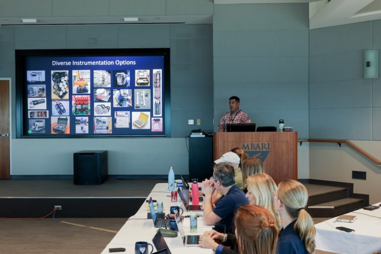 An engineer presents slides to an audience. The engineer has short black hair and is wearing a brightly patterned short-sleeved shirt. He is standing behind a wooden podium with the MBARI logo. Behind him is a projection screen displaying a presentation slide with several small photos titled, Diverse Instrumentation Options. In the foreground are workshop participants seated at a table draped in a white tablecloth. In the background are the gray walls of a conference room.