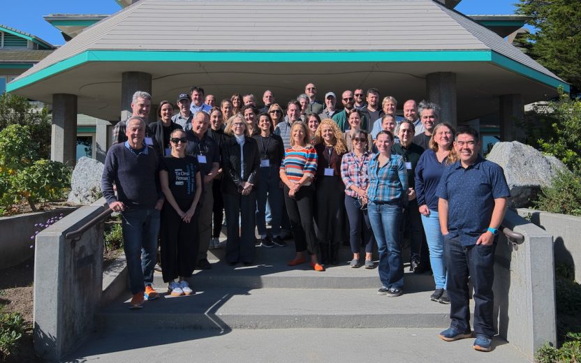 Thirty-seven workshop participants pose outdoors on the gray concrete steps. In the background is the gray roof and green trim of a building, green trees, and blue sky.