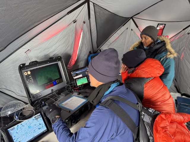Three engineers wearing cold-weather gear operate computer equipment controlling a robotic submersible deployed under the ice. The engineer in the foreground is piloting the submersible while the two engineers in the background monitor data feeds from the submersible. In the background is the silver lining of a tent.