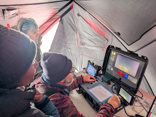 Three researchers wearing cold-weather gear operate computer equipment controlling a robotic submersible deployed under the ice. The engineer in the center is piloting the submersible while the two researchers on the left side of the frame observe the video feed from the submersible. In the background is the silver lining of a tent.