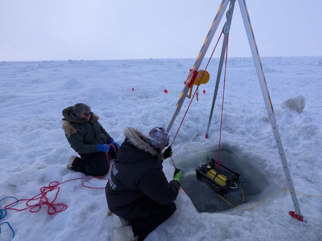 Two scientists wearing cold-weather gear retrieve a robotic submersible after deployment under the ice. The scientists are kneeling on white ice. On the right side of the frame is a silver metal tripod above a square hole cut into the ice. A pulley on the tripod holds a red rope tethered to the robotic submersible. The robotic submersible has a yellow metal casing and black metal frame. The background is a white ice shelf with light gray sky on the horizon.