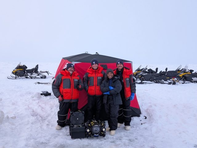 Four engineers wearing cold-weather gear pose for a photo with a robotic submersible in front of a red tent. In the foreground is white ice. In the background are several black-and-yellow snowmobiles and light gray sky.