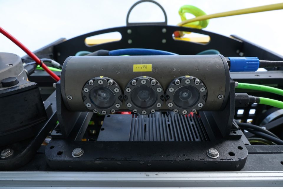 Photo documentation shows a camera system on a robotic submersible. The camera system has three round lenses and is housed inside a gray cylinder. In the background are assorted multi-colored cables.