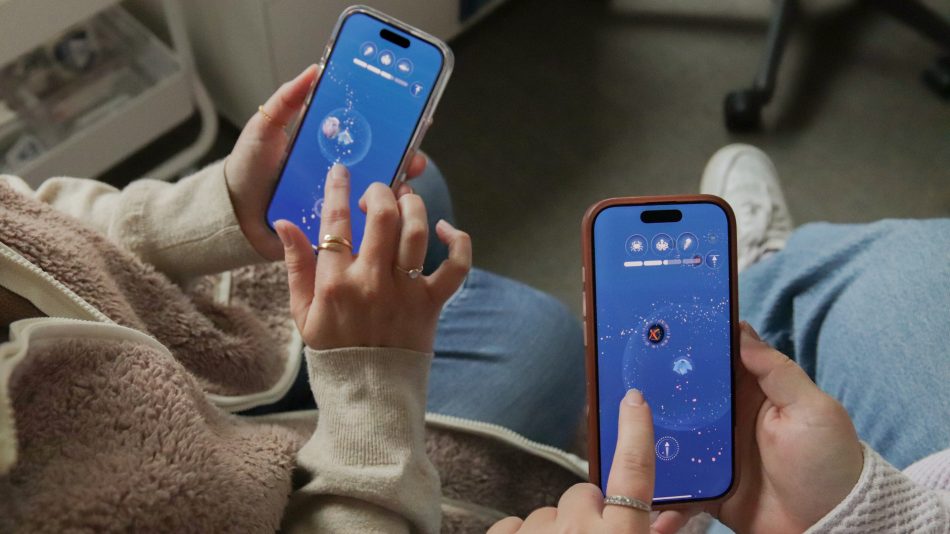 Two gamers seated on a couch play the FathomVerse mobile game on their iPhones. The gamer on the left is wearing a beige sweater, a fuzzy pale-pink vest, and blue jeans while holding an iPhone in a clear case. The gamer on the right is wearing a textured pale-pink shirt, blue jeans, and white sneakers while holding an iPhone in a pink case. Both iPhone screens are displaying an origami-like blue avatar moving through a stream of dots revealing circular photos of marine life, with three round animal icons and a progress bar at the top of the screen. Blurred in the background are a white cabinet, the black plastic leg of a chair, and gray carpet.