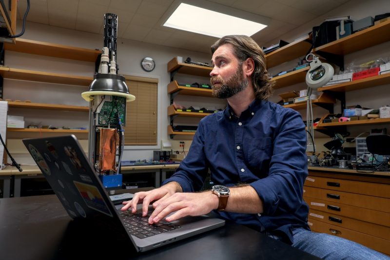 A scientist types at a computer while seated behind a black workbench in an engineering laboratory. He is looking at the cylindrical, black, plastic sensory array from a robotic float. In the background are wooden drawers and cabinets with assorted pieces of engineering equipment.