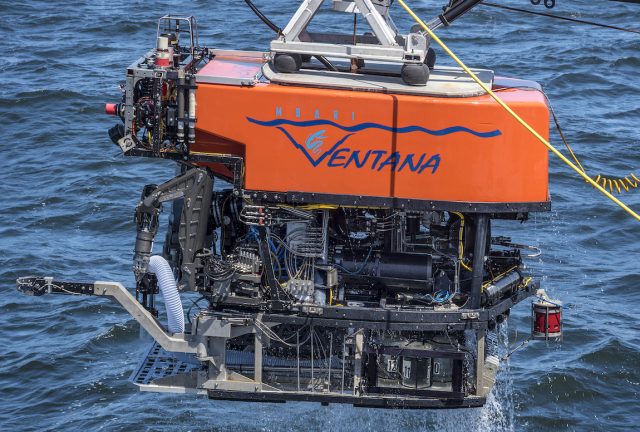 An MBARI remotely operated vehicle being lifted out of the water. The robotic submersible has a bright orange float with the name “Ventana” with MBARI’s gulper eel logo serving as the “V.” Beneath the float is a black metal frame and various pieces of equipment and wires, including a black-and-silver robotic arm and a transparent hose. Seawater is dripping off the vehicle and blue ocean is visible in the background.