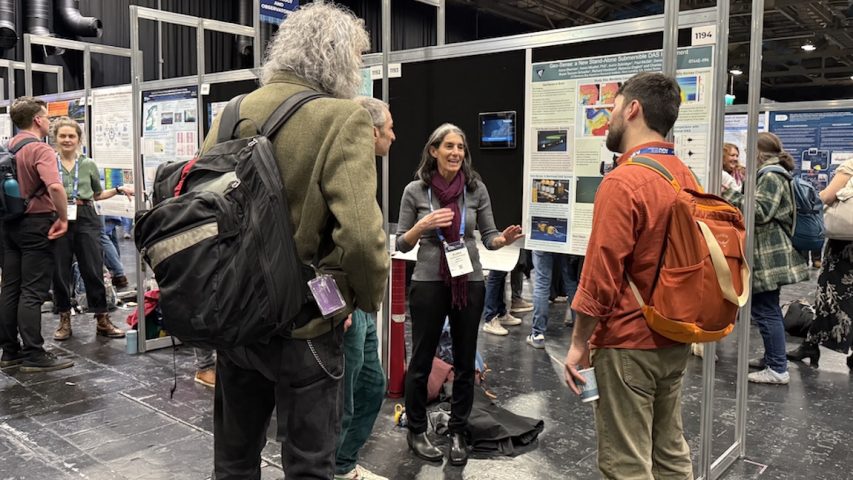 An engineer speaks with attendees at a scientific conference. The engineer has long black hair with a gray streak and is wearing a maroon scarf, a gray sweater, black pants, and black boots. She is standing in front of a scientific poster mounted to a black board and speaking to three conference attendees who have their backs to the camera. In the background is the crowded conference hall filled with attendees discussing scientific posters.