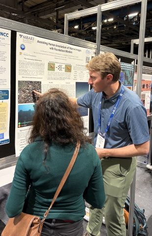 A software engineer speaks with an attendee at a scientific conference. The engineer has short blond hair and is wearing a blue lanyard identification badge, blue polo shirt, and green pants. He is standing in front of a scientific poster mounted to a black board and speaking to a conference attendee with long brown hair and wearing a teal sweater, gray pants, and a tan purse who has her back to the camera. In the background are more boards with scientific posters.