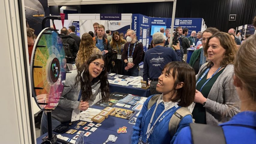 An executive coordinator watches visitors spin a wheel for a prize at a conference exhibitor booth. The executive coordinator has long black hair and is wearing glasses, a blue lanyard, a gray sweatshirt, and blue jeans. She is on the left side of the frame and is leaning over a table, watching a colorful spinning wheel labeled with prize items. On the right side of the frame are several conference attendees. In the background is a busy conference hall with tables, posters, and crowds of conference attendees.
