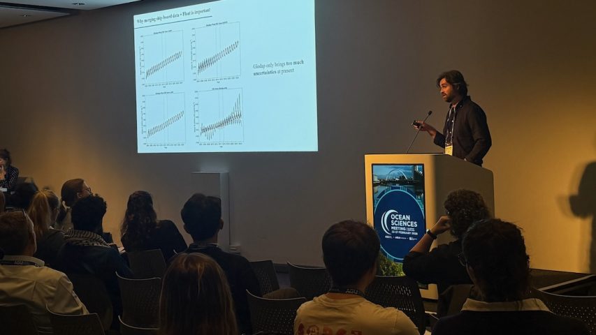 A scientist presents at a scientific conference in a darkened room. The scientist has short, wavy, brown hair and is wearing a blue shirt. He is standing behind a white podium displaying the logo for the Ocean Sciences Meeting 2026. In the foreground is an audience of attendees seated in beige chairs. In the background is a white wall with a presentation slide projected on it.