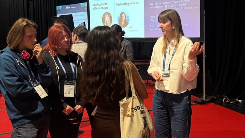 An engineer speaks with attendees at a town hall at a scientific conference. The engineer has long blonde hair and is wearing a blue lanyard, a white shirt, and blue jeans. She is standing in front of a projector screen displaying a presentation slide and is speaking to three conference attendees. In the background is the crowded conference hall filled with attendees.