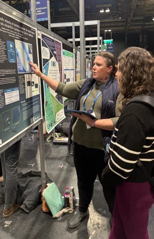 A research technician speaks with an attendee at a scientific conference. The research technician has long brown hair, is wearing a blue lanyard, a navy blue vest, a green sweater, black pants, and black boots, and is holding a tablet computer. She is standing in front of a scientific poster mounted to a black board and speaking to a conference attendee who has their back to the camera. In the background is a conference hall filled with scientific posters.