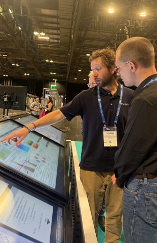 A postdoctoral fellow speaks with an attendee at a scientific conference. The engineer has short blond hair and a short blond beard and is wearing a blue lanyard identification badge, navy blue polo shirt, and tan pants. He is standing over a large television monitor displaying a scientific poster and is speaking to a conference attendee while pointing to something on screen with his right hand. In the background is a conference hall with attendees and scientific posters.