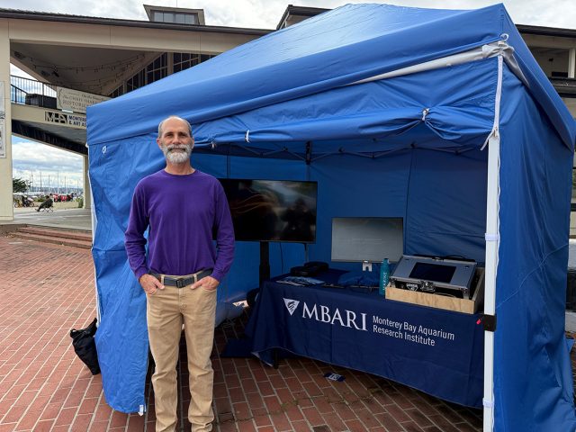 A scientist wearing a purple shirt and tan pants stands in front of a bright blue event tent. A table inside the tent has a large monitor screen displaying underwater video and an interactive monitor and speaker. In the background is an outdoor red-brick plaza with a tan two-story building, and behind it a wharf and small boats.