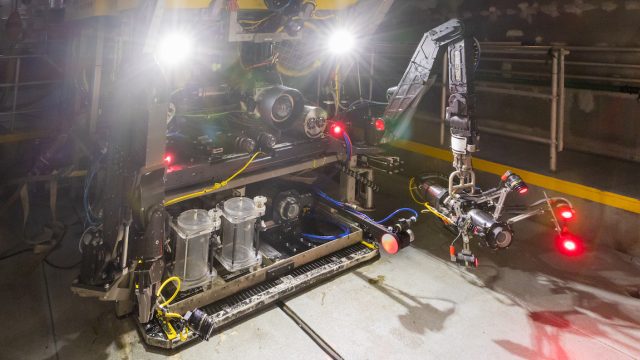 A deep-sea robot with a yellow float at the top, a black metal frame in the middle, a silver metal frame, and two transparent acrylic cylinders at the base sit on the gray deck of a research vessel. Two bright white headlights are shining at the top of the robot. A silver metal mechanical arm is on the left, and two camera systems are on the right. The camera system in the foreground has a black silver canister on a black metal rod that shines a red light. A silver mechanical arm holds the camera system in the background and has a gray metal canister with five red lights. This robot was photographed indoors, from the side, sitting on a pair of mechanical doors with various maritime equipment visible in the background.