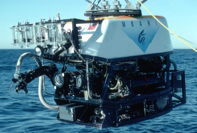 An MBARI remotely operated vehicle being held out over the water for deployment. The robotic submersible has a white float with the name “MBARI” and MBARI’s gulper eel logo printed in dark blue above the V-shaped submarine canyon outline filled in a light blue gradient. Four transparent cylinders are at the front of the float. Beneath the float is a black metal frame and various pieces of equipment, including a black camera, a black metal robotic arm, and a white hose. Grayish-blue ocean and blue sky are visible in the background.