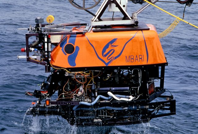 An MBARI remotely operated vehicle being lifted out of the water. The robotic submersible has a bright orange float with three blue marks at the front (left of photo) and MBARI’s gulper eel logo and the name “MBARI” printed in dark blue at the center. Beneath the float is a black metal frame and various pieces of equipment, hoses, and wires. Seawater is dripping off the vehicle and grayish-blue ocean is visible in the background.
